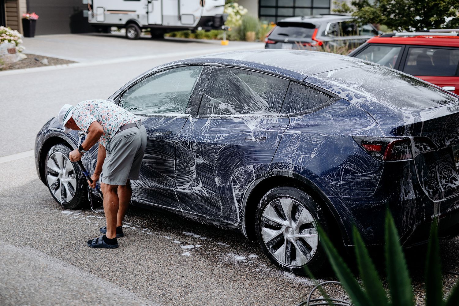 A bride's father washes a dark blue Tesla Model 3 sedan in a parking lot using soap and water.