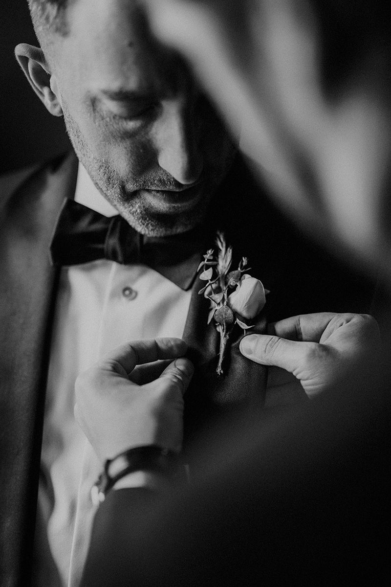 Black and white close-up photo showing a man adjusting his bowtie and another man fixing his boutonniere.
