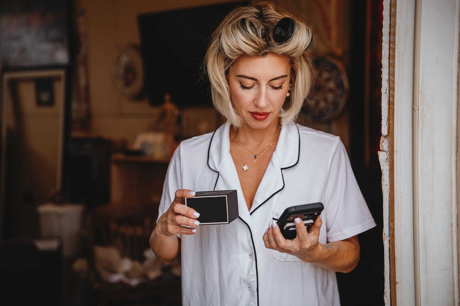 A bride in a white robe enjoys a coffee while her hair sets within large rollers.