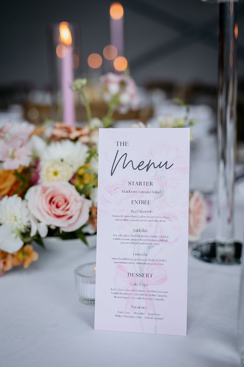 Elegant wedding reception menu cards and floral centerpieces with pink roses on white tablecloths in romantic lighting.