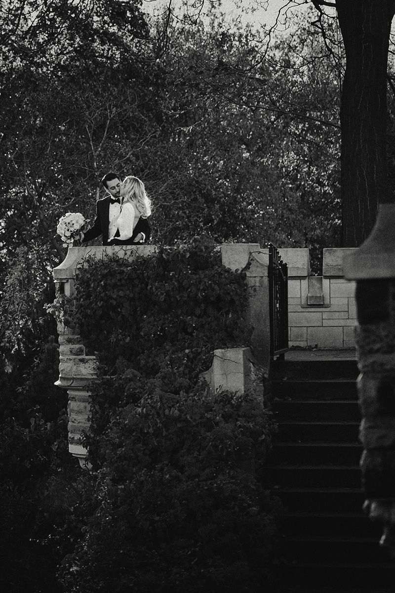 A black and white photograph of a couple standing on stone steps surrounded by lush garden foliage.