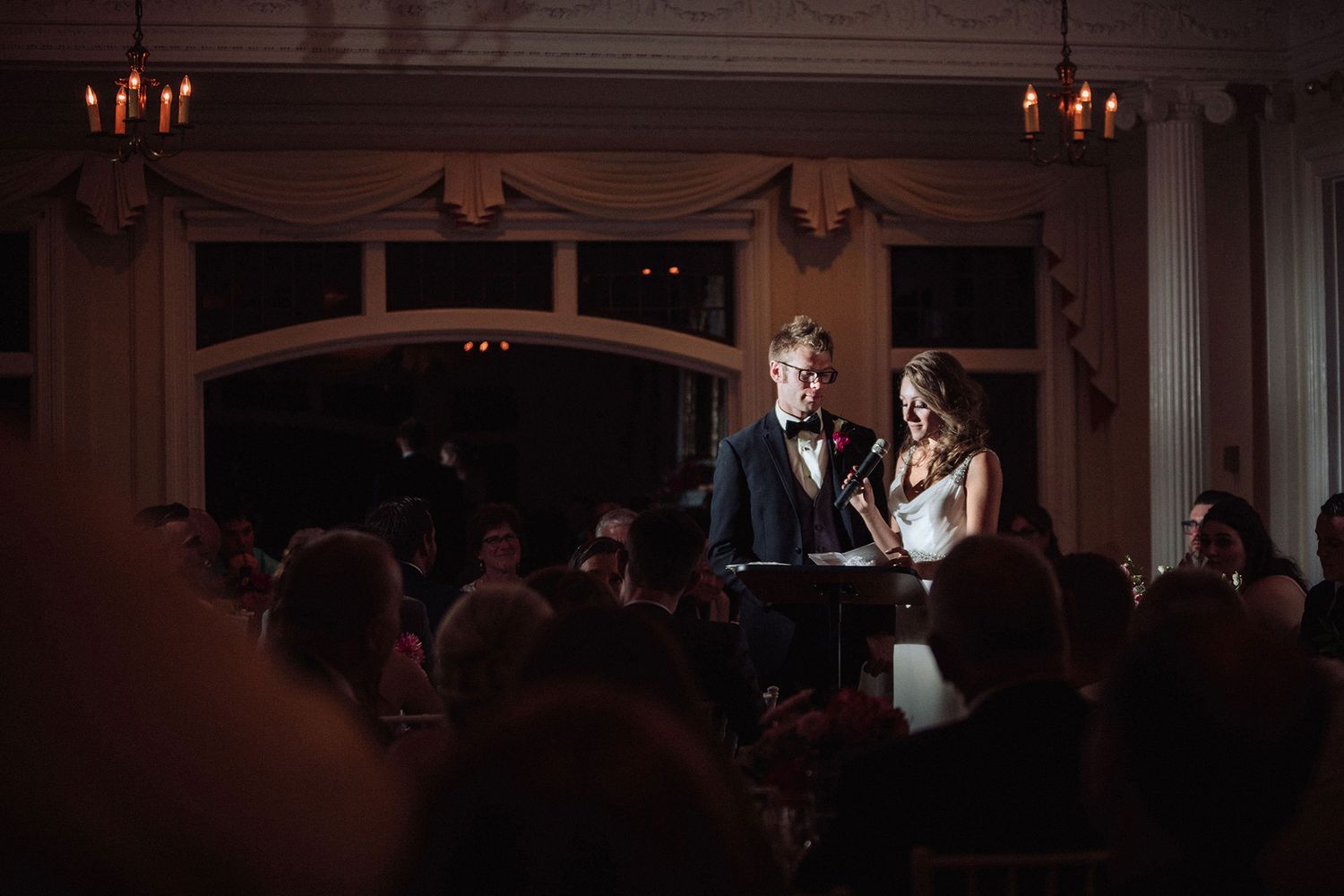 A dimly lit wedding reception venue with a spot light on a bride and groom as they give a speech at a lectern.