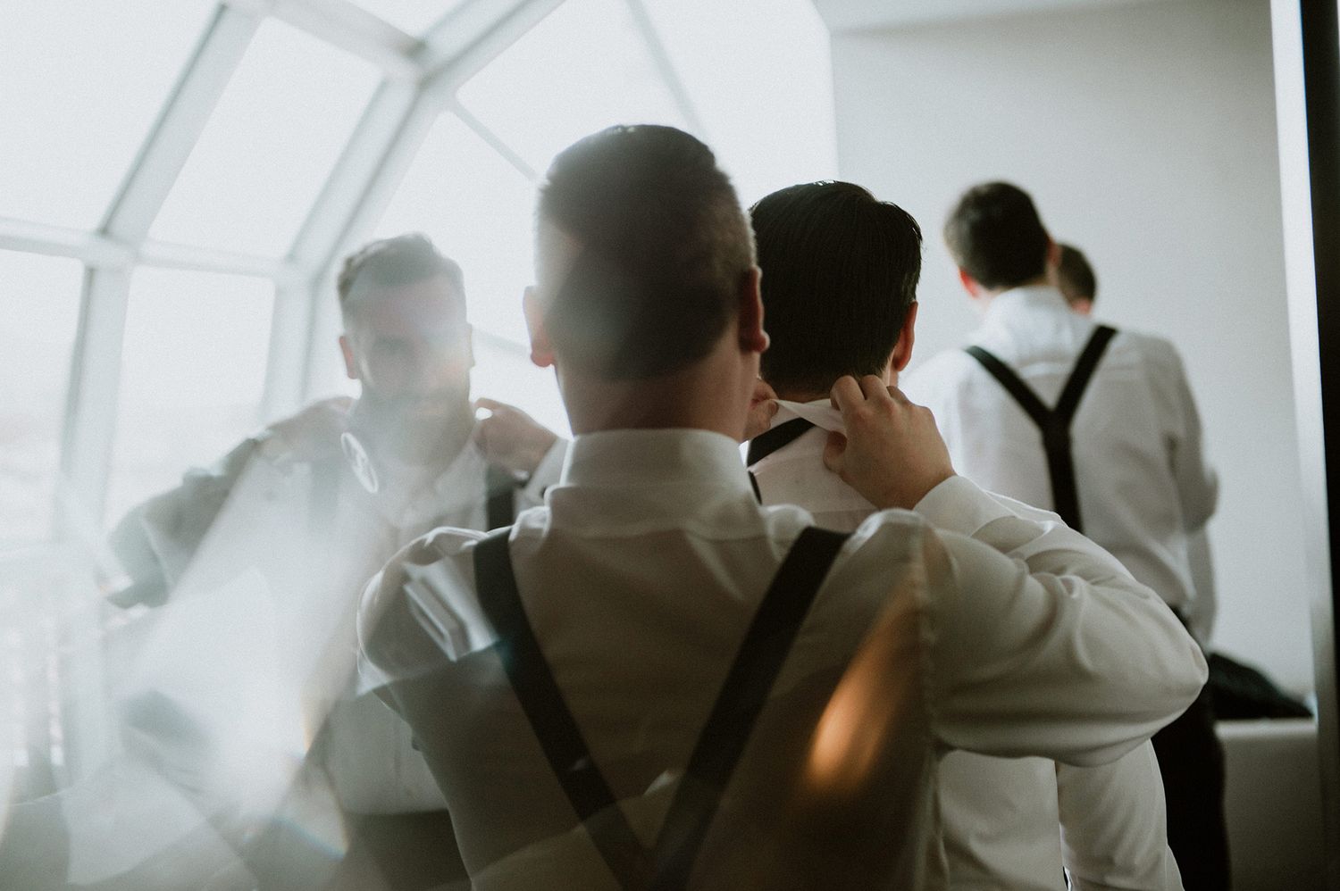 A group of grooms men stand in a bright, art-deco space looking out large windows as they help each other with suspenders.