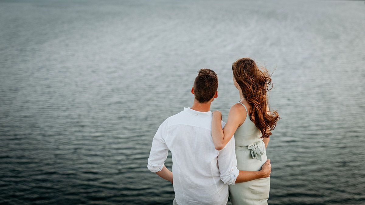 Couple standing together embracing while gazing at a calm lake, viewed from behind in romantic outdoor setting.