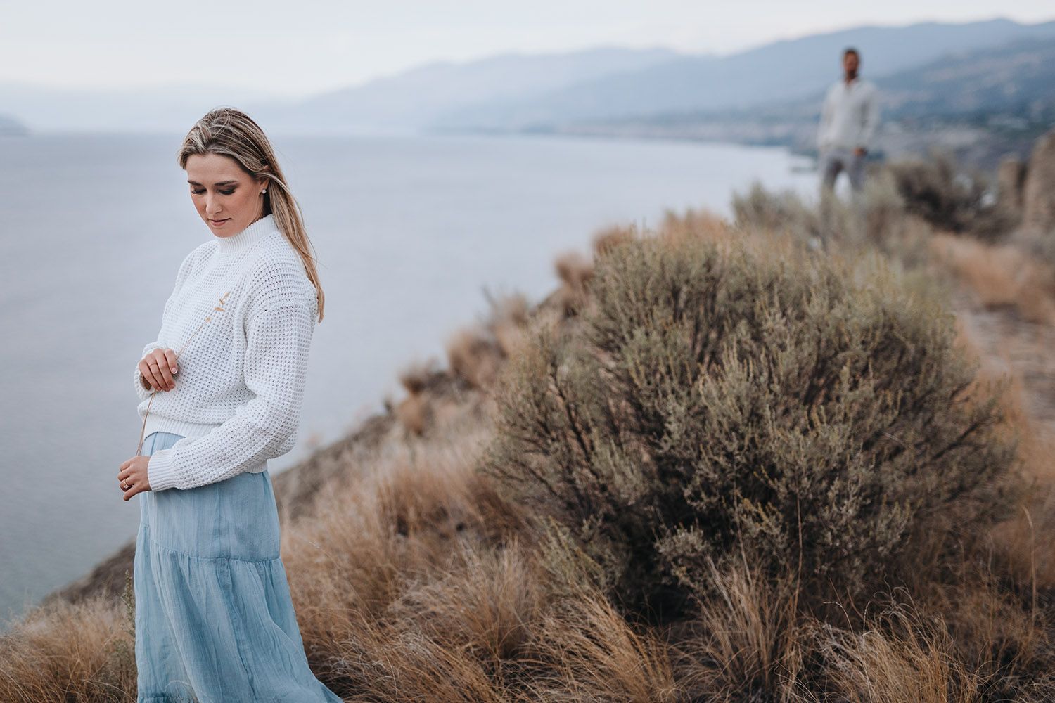 Woman in white sweater and blue dress walking through golden mountain meadow at sunset with scenic lake Okanagan in the background.