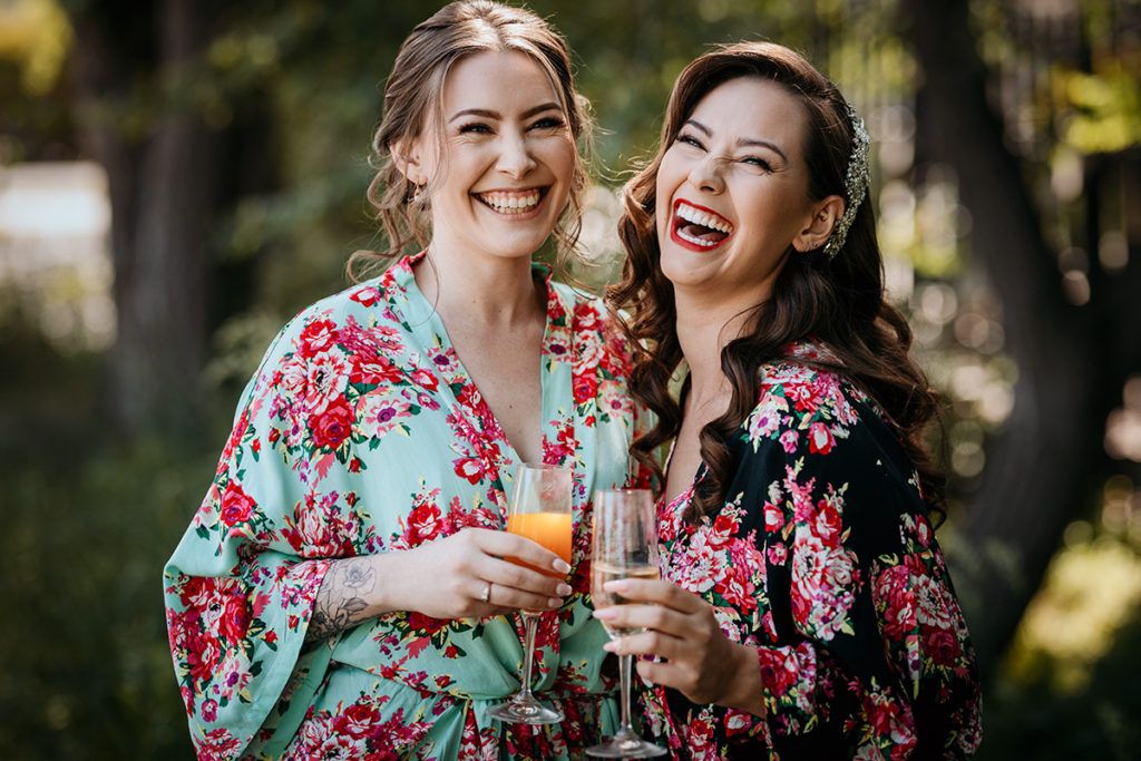 A bride and maid of honour in floral robes share a joyful moment while holding drinks outdoors in a garden setting.