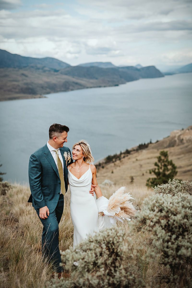 A couple in wedding attire embraces on a scenic hillside overlooking a serene mountain lake on their special day.