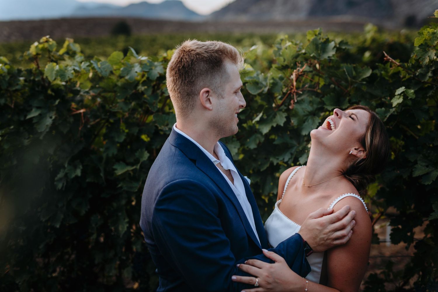 A couple shares a joyful moment together while standing in a vineyard with mountains in the background. The bride is laughing up at the sky.