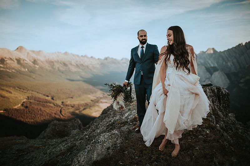 Couple in wedding attire stands together on scenic mountain peaks during golden sunset hour.