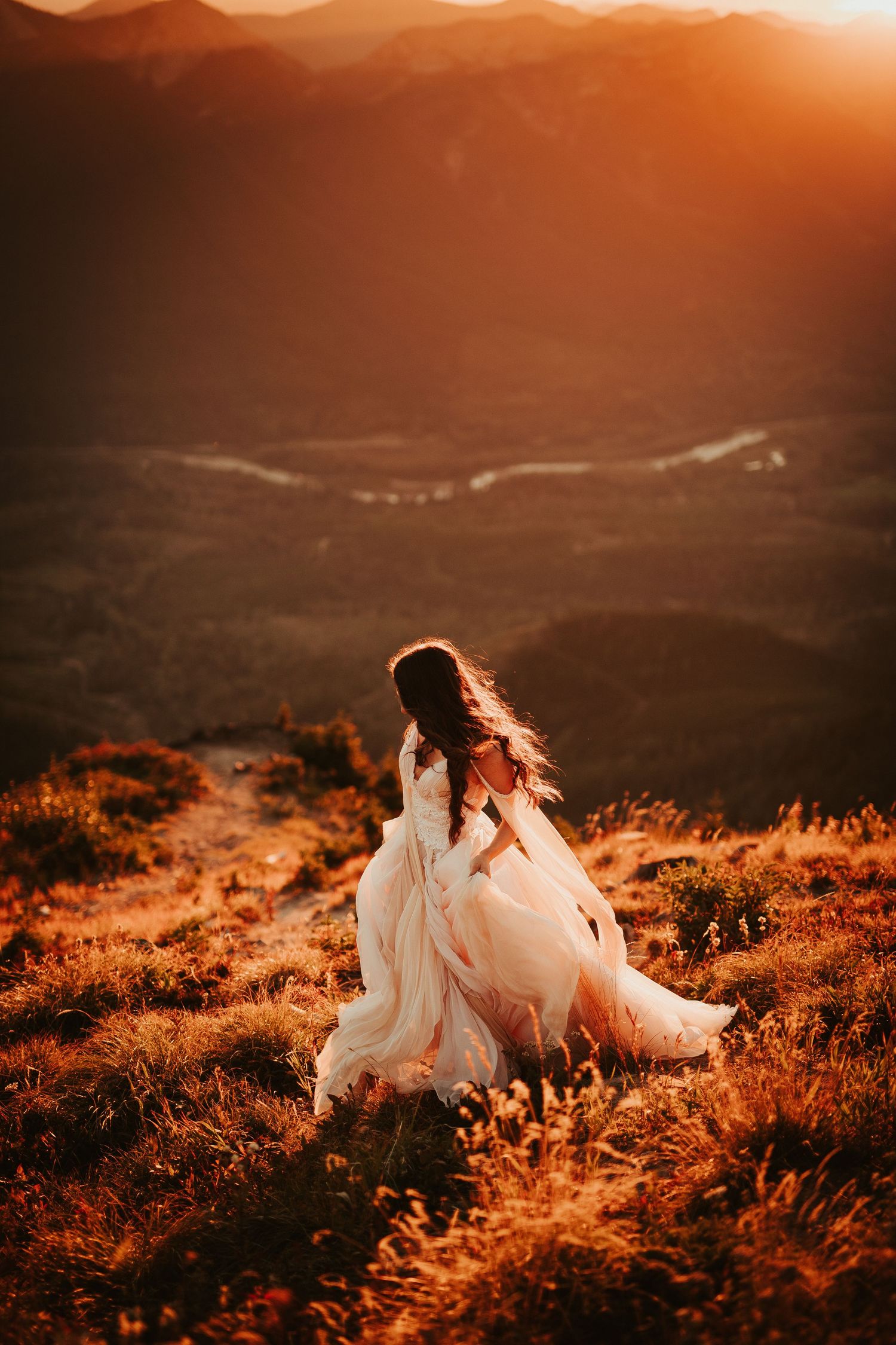 A bride on a hilltop at sunset with dramatic orange lighting and mountain views.