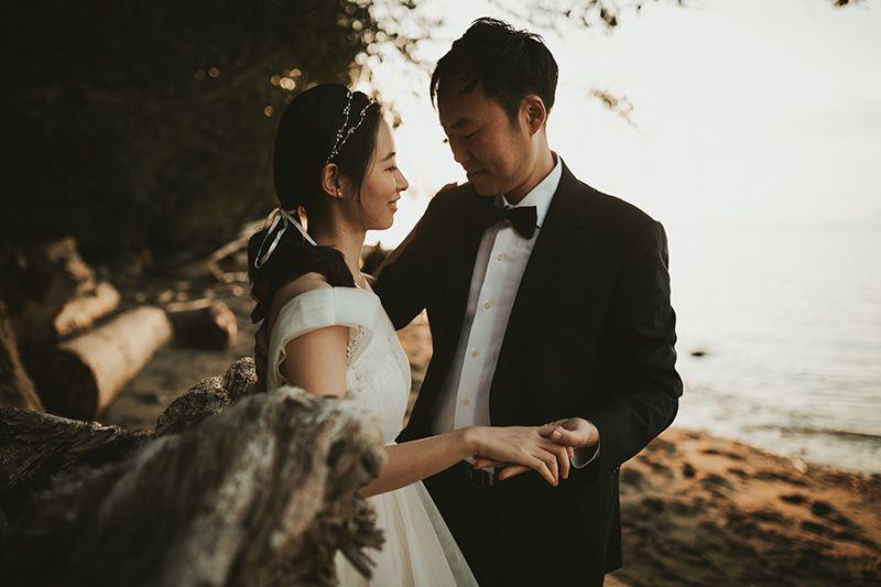 A romantic couple in wedding attire shares an intimate moment at sunset on a beach with dramatic lighting.