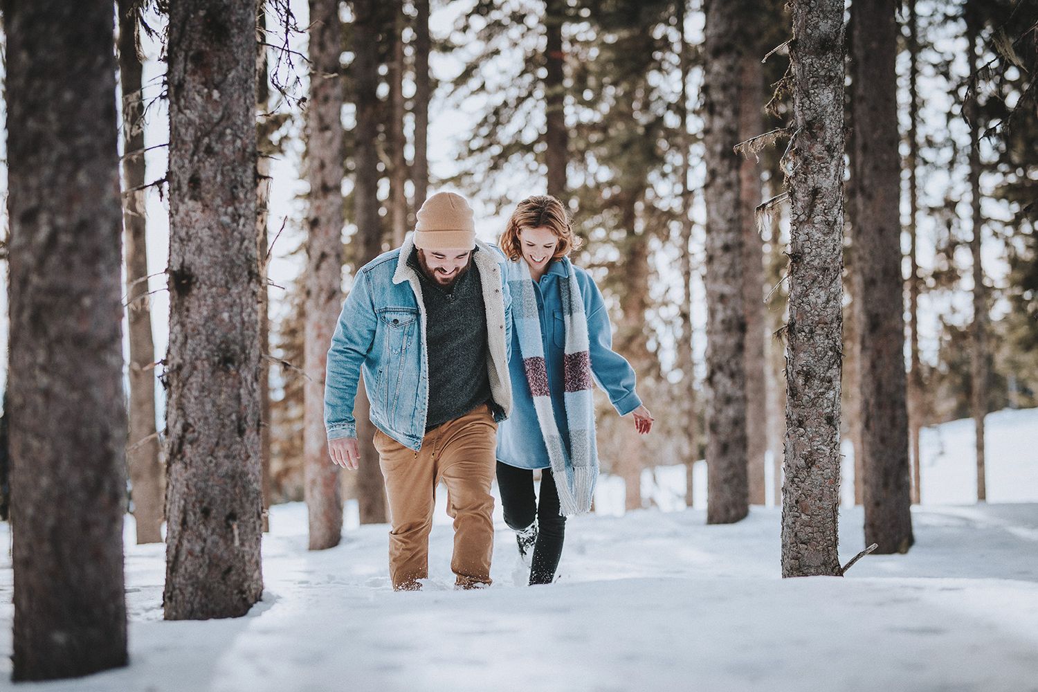 A couple in matching denim jackets walks through a snowy forest path on a winter day.