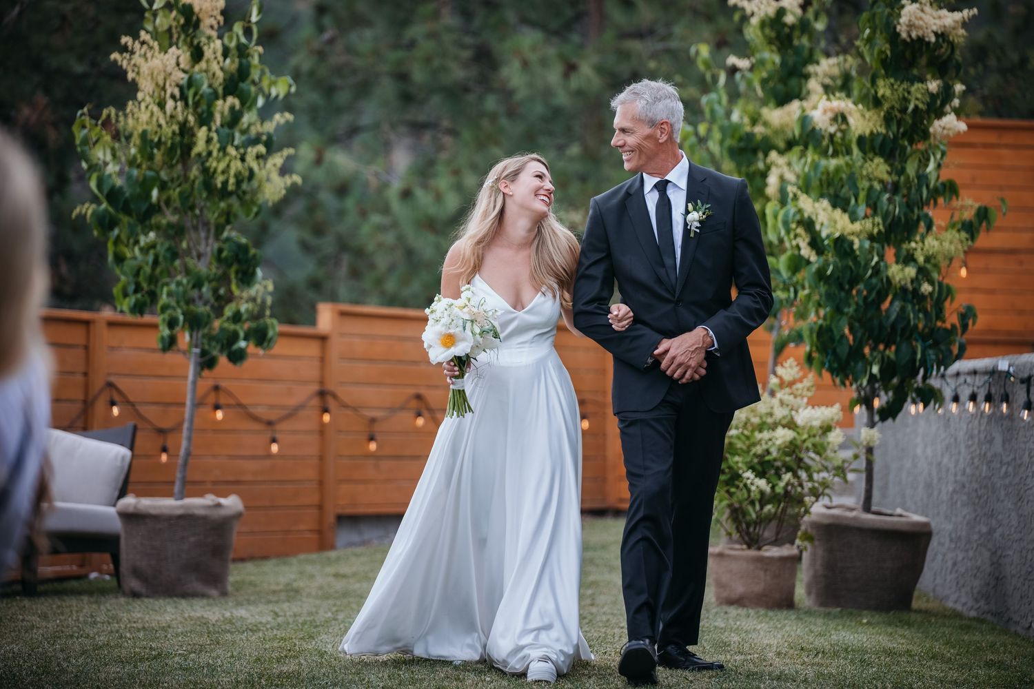 A bride and her father walk the aisle at a romantic outdoor wedding scene with string lights and wooden planters in a garden setting.
