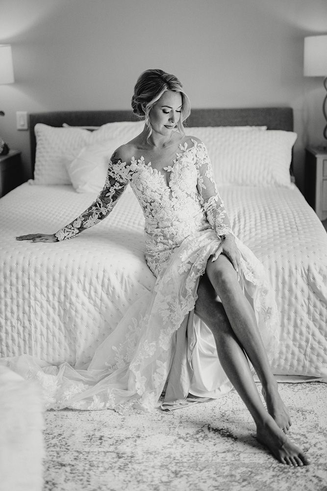 A bride in a lace wedding gown lounges gracefully on a white bed in a romantic black and white photo.