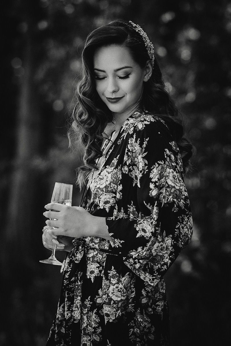 Black and white portrait of a bride-to-be in floral robe holding a champagne glass against bokeh background.