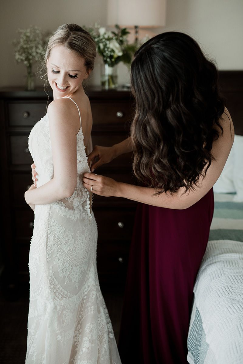 A bridesmaid in burgundy helps button up a delicate lace wedding dress during wedding preparations.