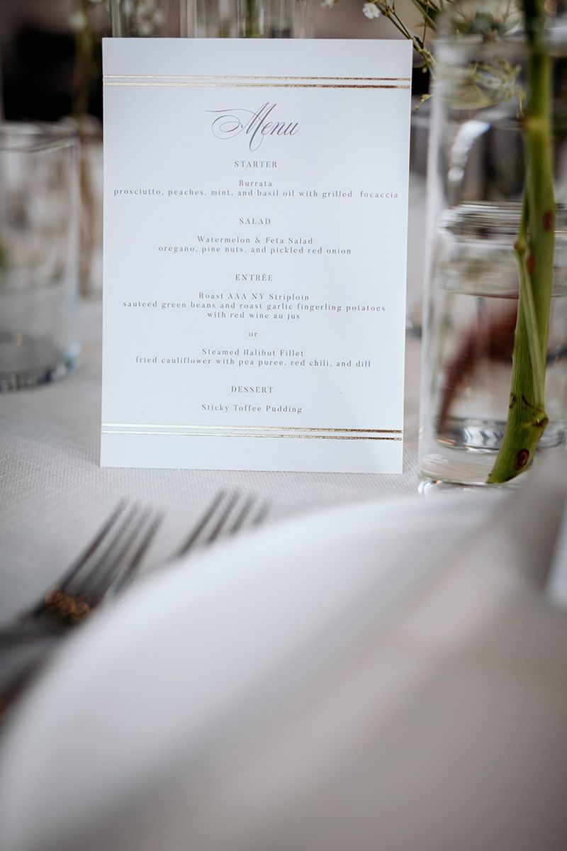 An elegant wedding menu card with gold accents stands on a white tablecloth next to silverware and glassware.