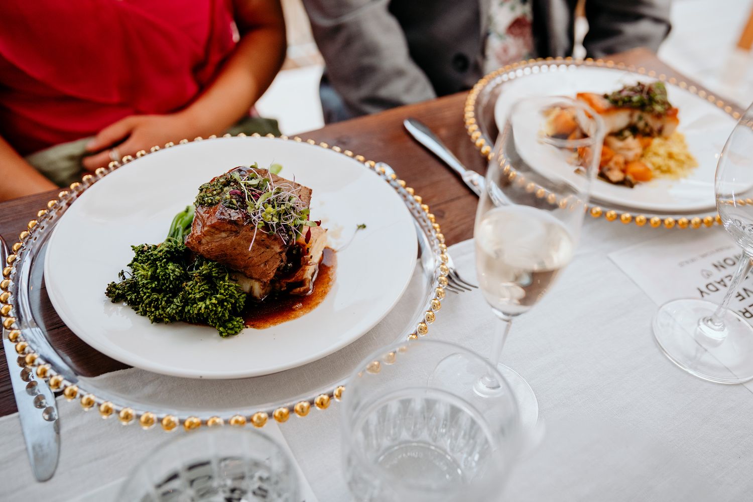 An elegant plated dinner with meat and sauteed greens served on gold-rimmed plates with wine glasses on a white tablecloth. Vows and Vines.
