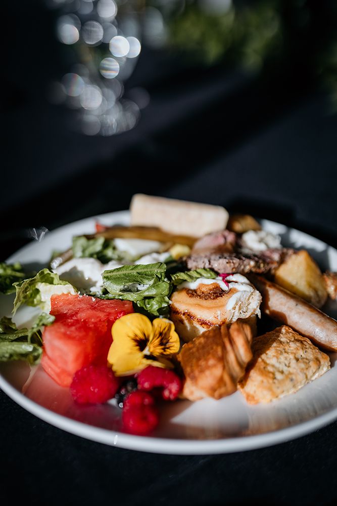 A colourful plate of food featuring watermelon, edible flowers, salad greens, and various cooked items arranged artfully.