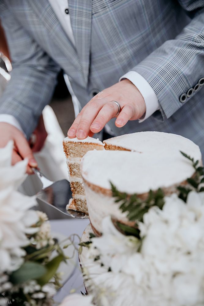 Close-up view of hands holding a slice of wedding cake with white flowers and greenery in the background.