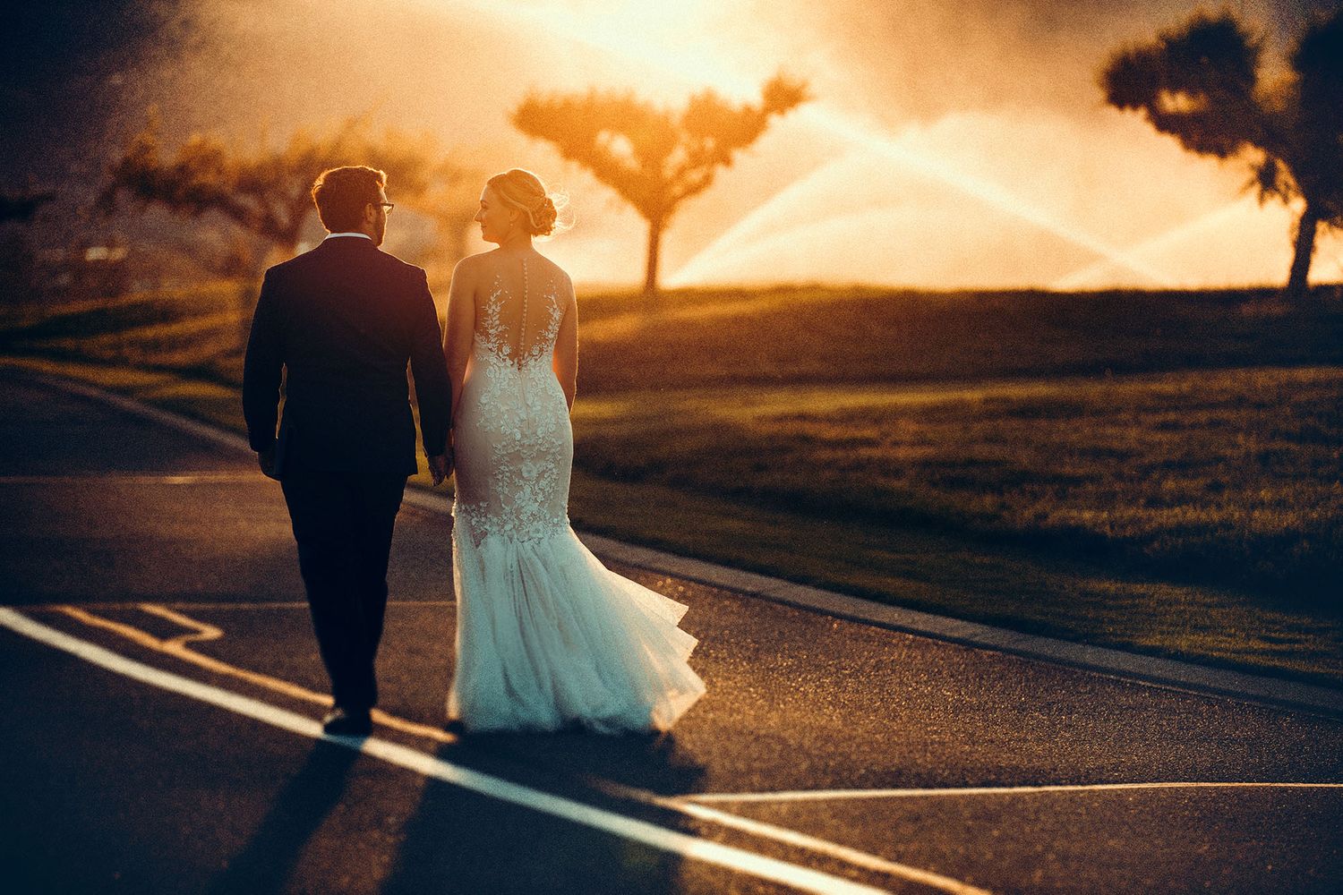 Stunning sunset photo of a bride and groom walking on a road with sprinklers watering the lawns at The Harvest Golf Club in Kelowna, BC.