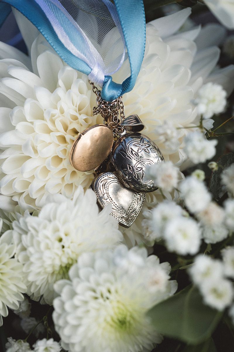 Vintage silver locket pendant with blue ribbon nestled among delicate white chrysanthemum flowers in a soft focus setting.