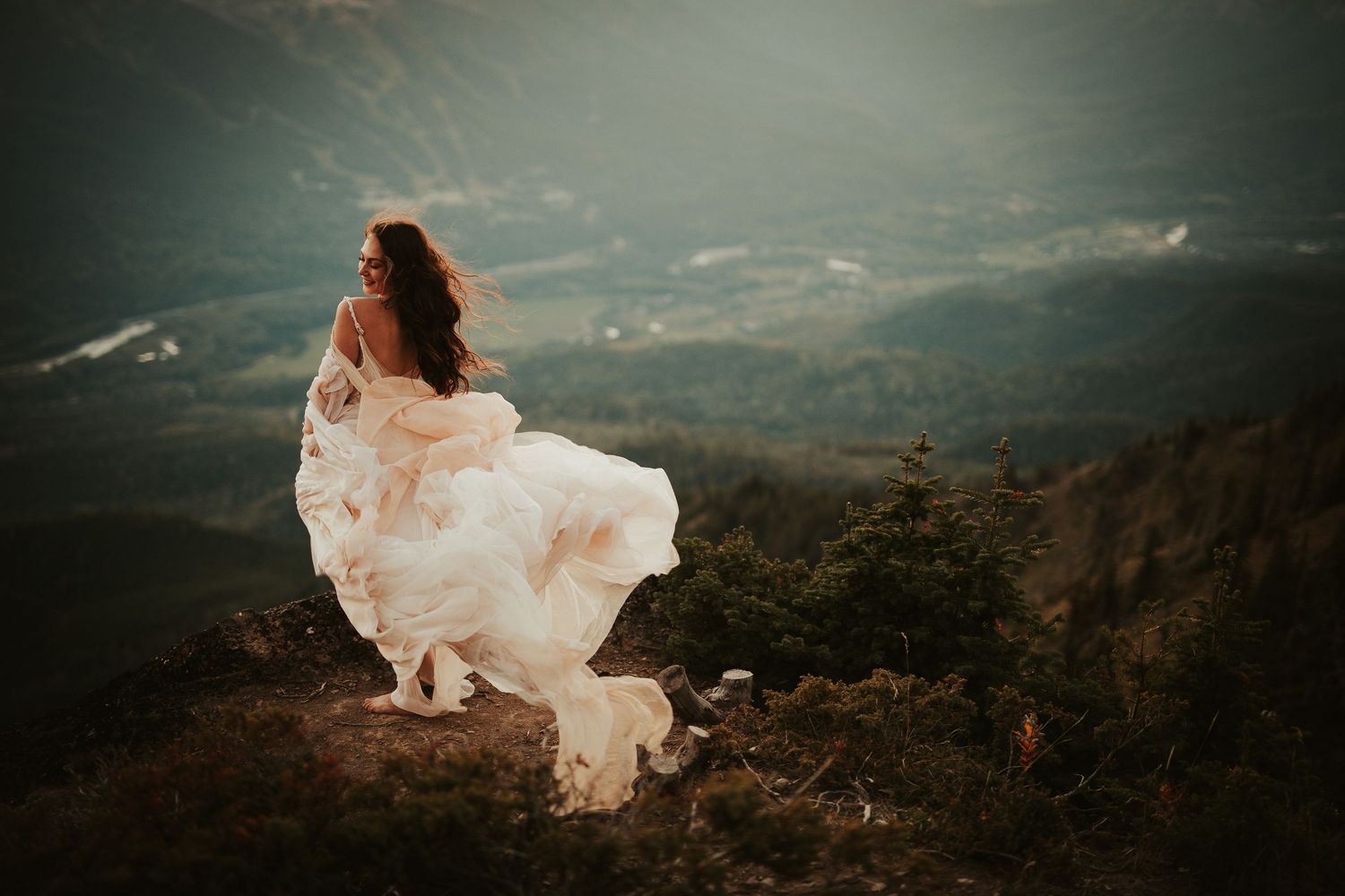 A bride in a flowing white dress that billows dramatically against a misty mountain landscape at sunset.