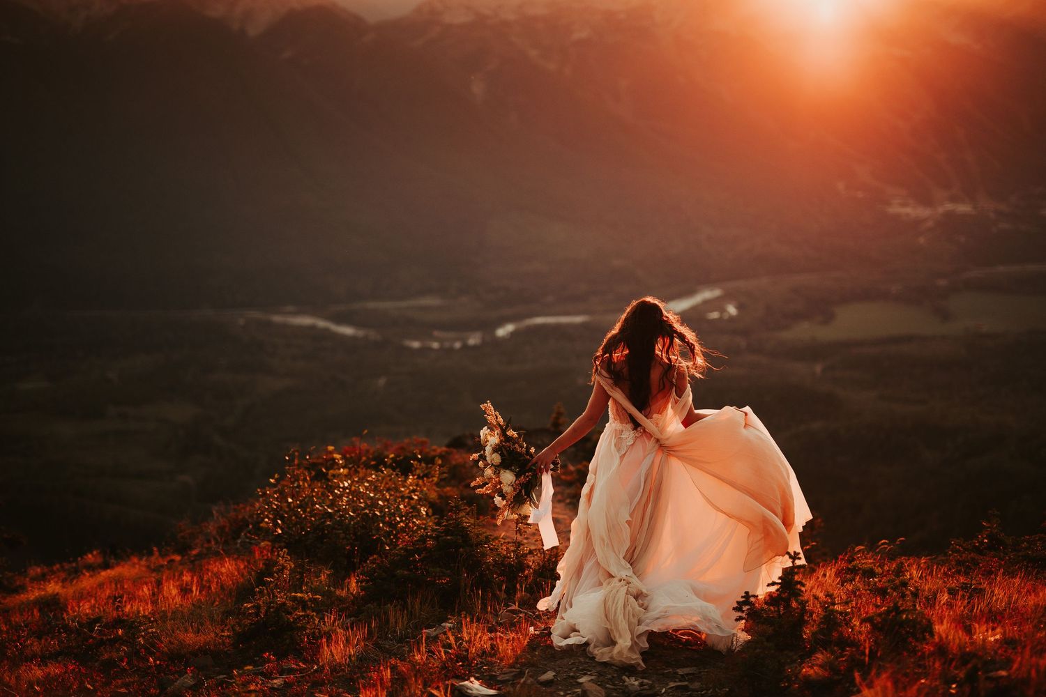 A romantic sunset wedding photo shoot silhouettes a bride against an orange sky in a mountain landscape.