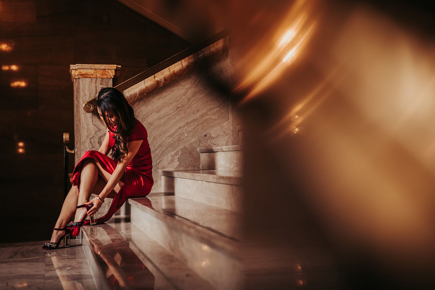 A Chinese bride in a red dress and heels sits on the marble steps of Hotel Vancouver in a moody, atmospheric setting.