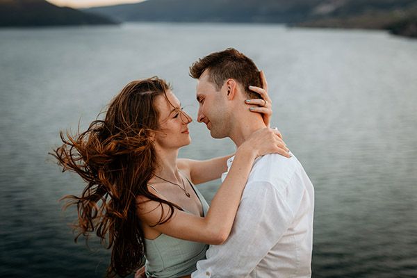 Romantic couple embracing by a serene lake at sunset, woman's hair blowing in the wind.