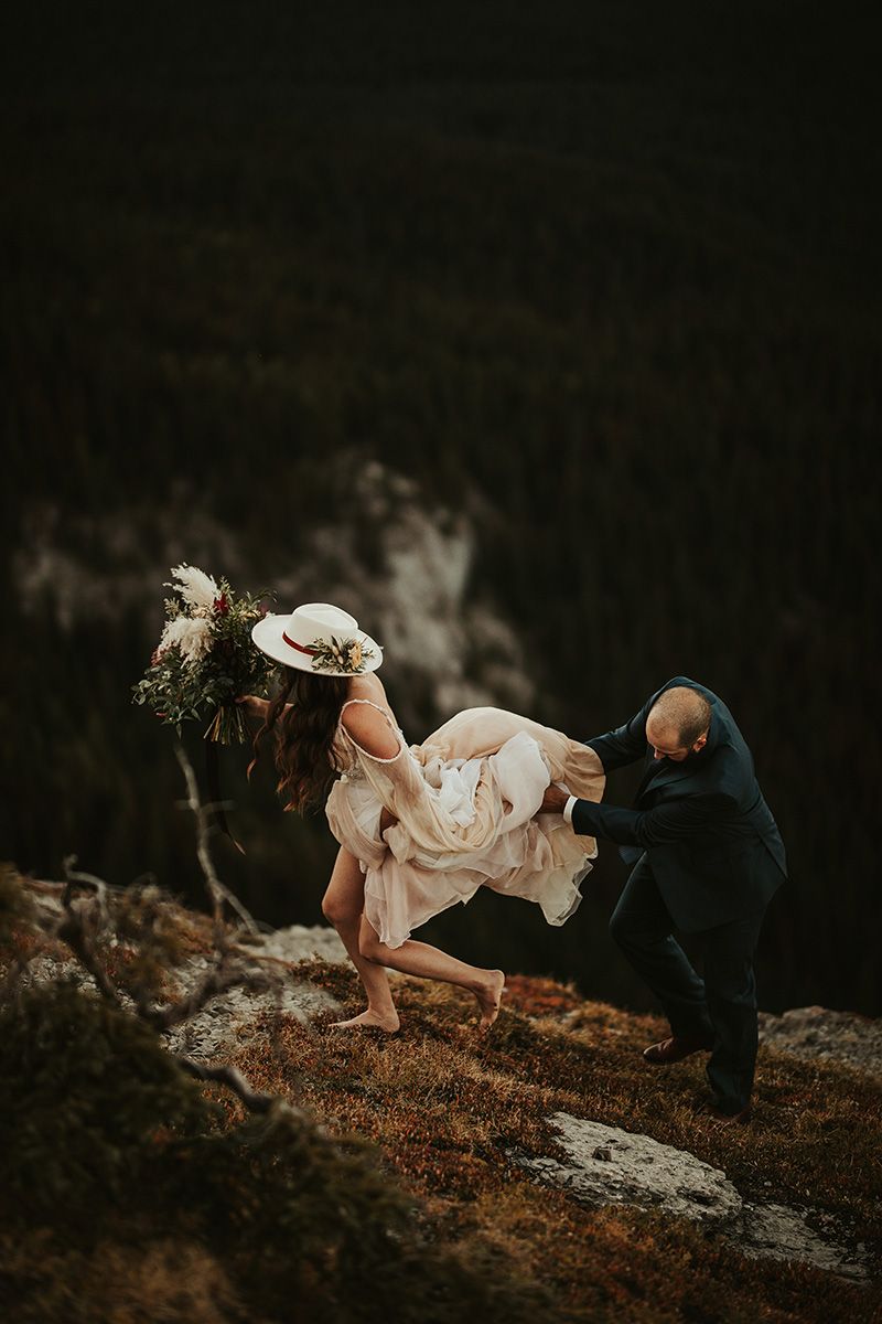 A groom holds his bride's dress as they both negotiate a a cliff edge in bare feet during a wedding photo shoot.
