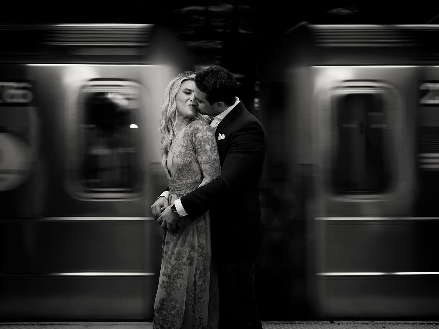 Motion blur of subway train cars passing through station platform while a romantic couple embrace in the foreground in black and white.