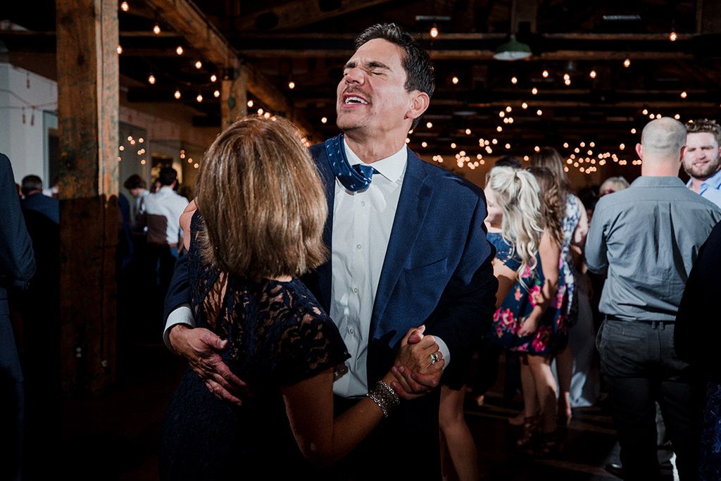A couple dances together at an indoor reception venue with exposed wooden beams and string lights overhead.