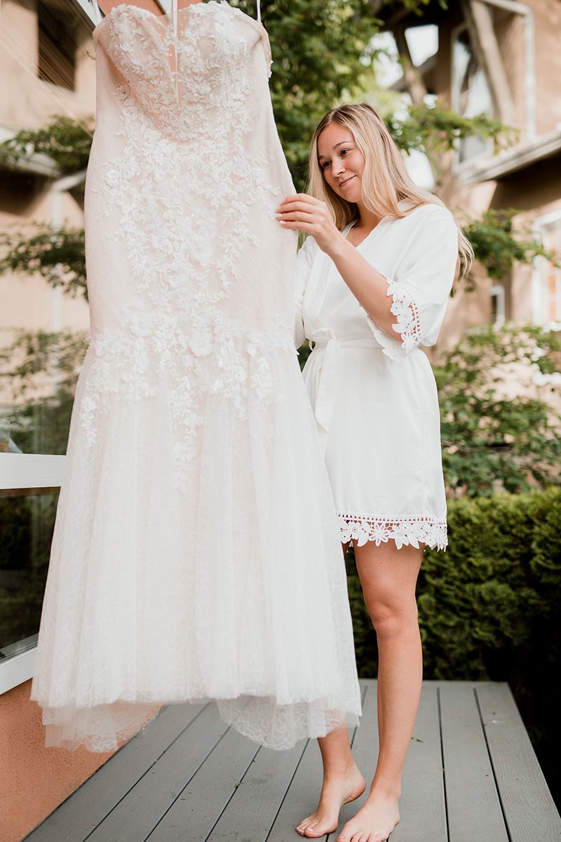 A bride-to-be inspects the details of a white lace wedding dress and bare feet on a wooden deck.