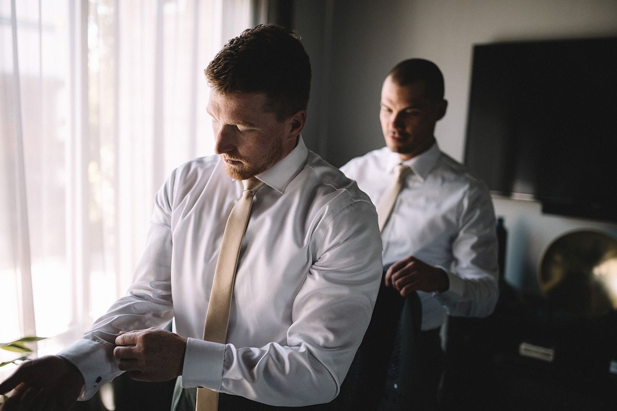 A man in a white dress shirt and pink neck-tie, adjusts his shirt cuff while his friend looks on.