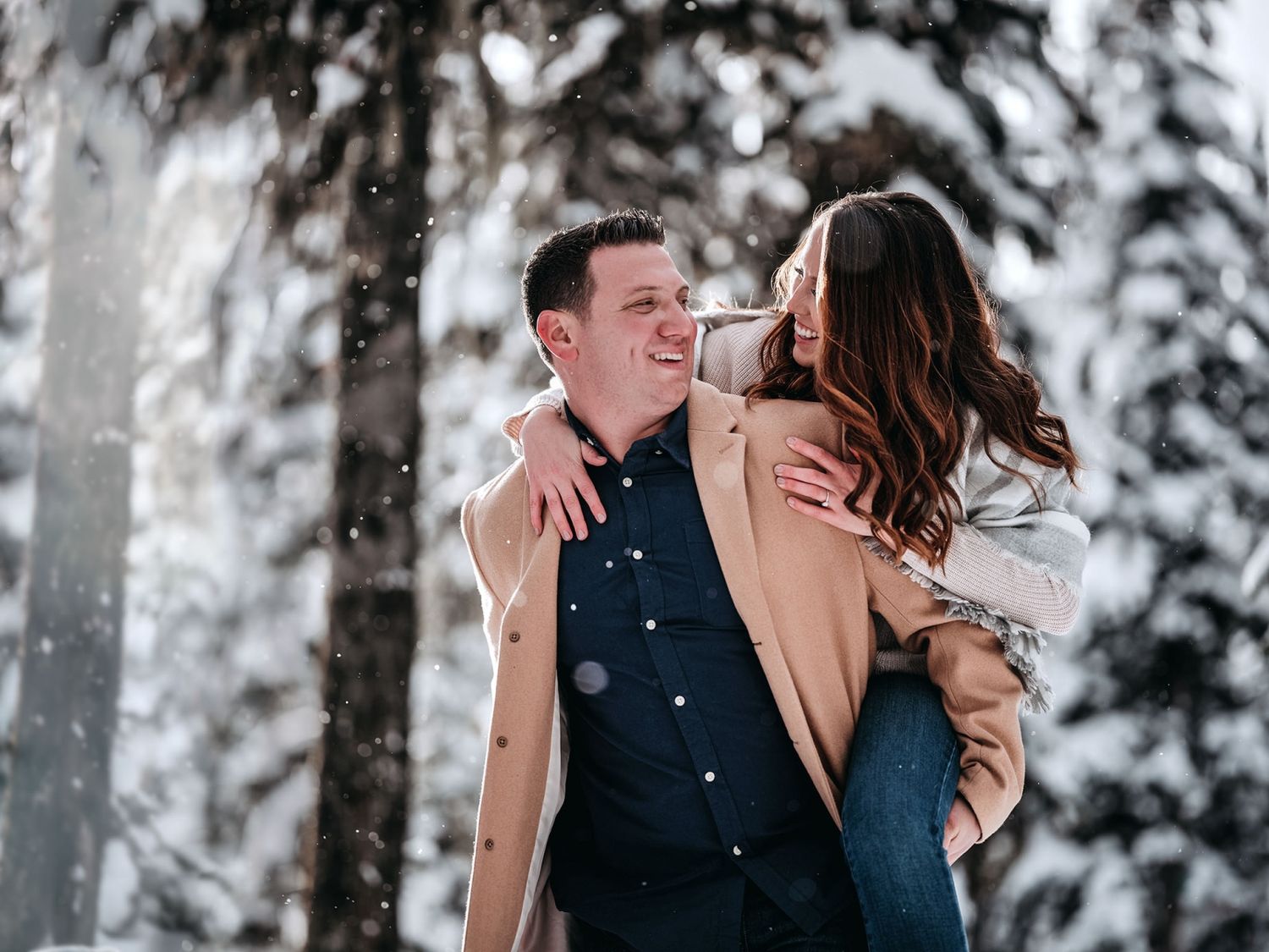 Romantic couple embraces in a snowy winter forest with snow-covered evergreen trees in the background.