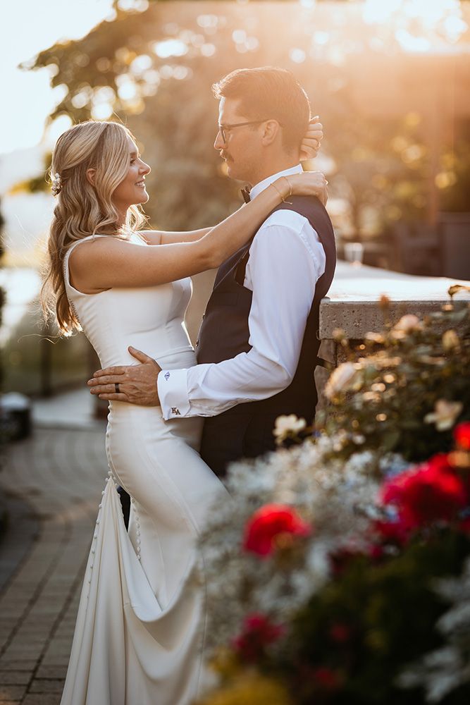 Newlywed couple embraces during golden hour sunset with red flowers in foreground at outdoor garden venue.