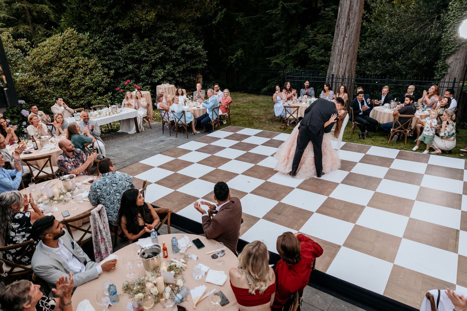 Wedding guests sitting at their tables surrounding a checkered dance floor, applaud a bride and groom at an outdoor reception celebration.