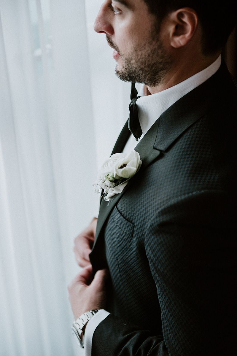 Close-up view of groom adjusting black tie and boutonniere against white curtain background.