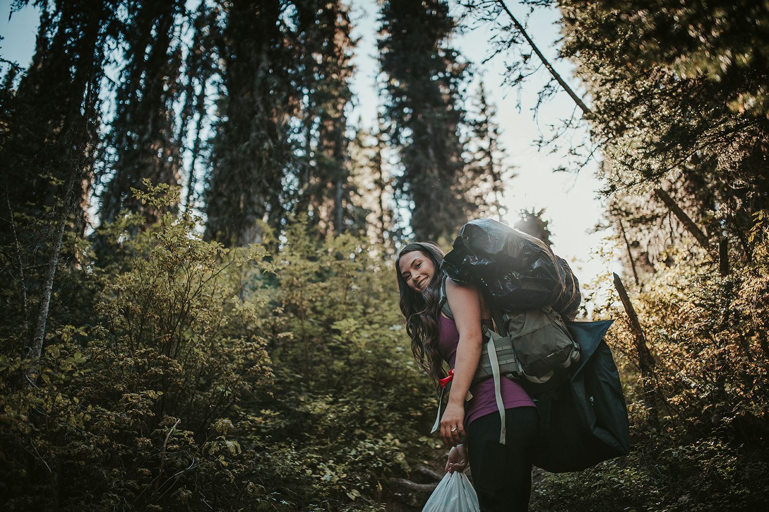 A hiker with a backpack bends down to pick up trash in a sunlit forest trail.