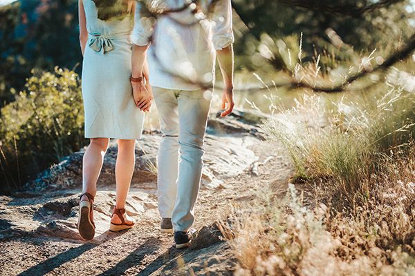 Couple holding hands on a scenic rocky trail surrounded by golden grasses and trees in warm sunlight.