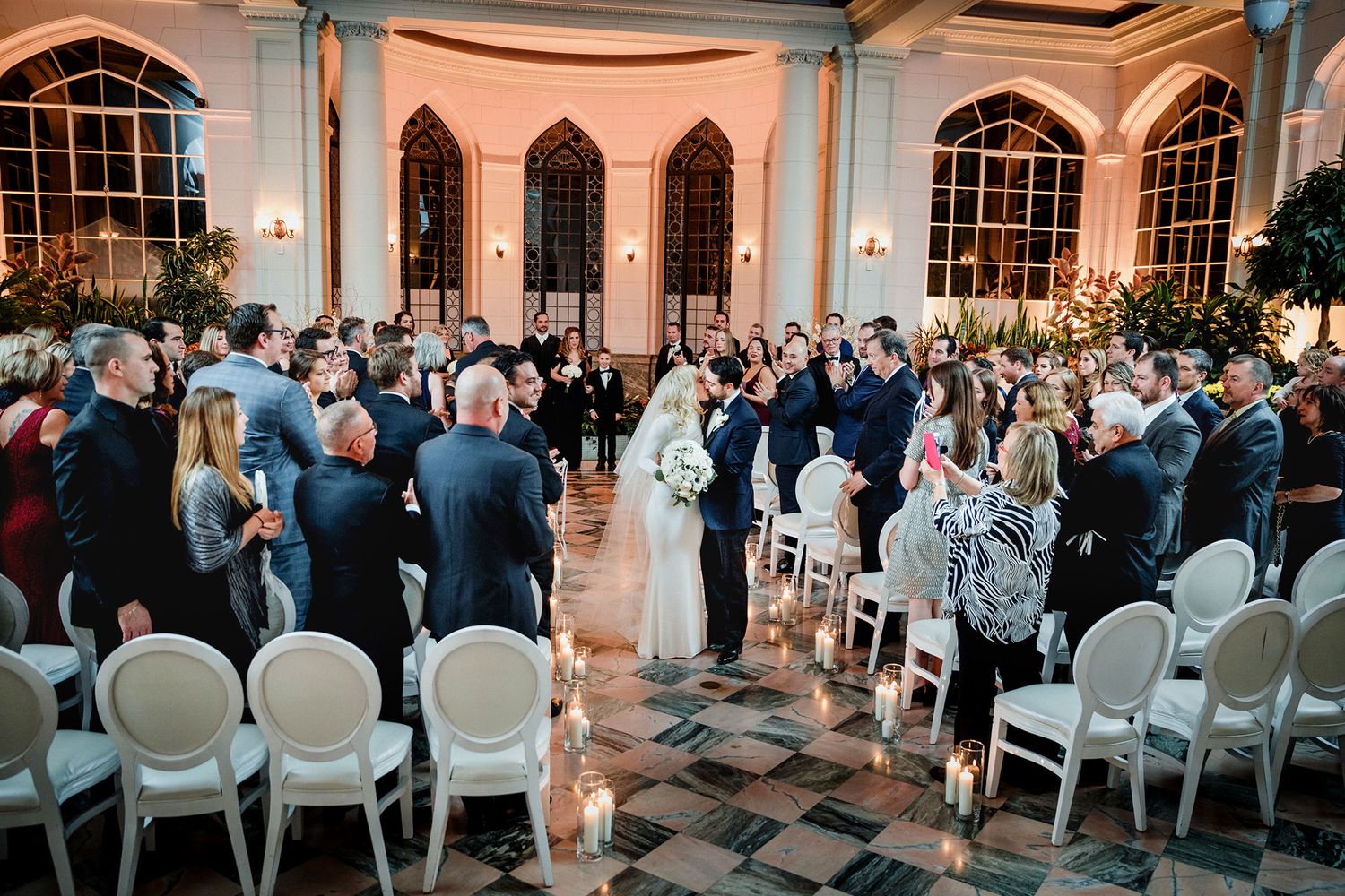 Wedding guests stand and watch as a couple shares their first kiss in an elegant ballroom with arched windows.