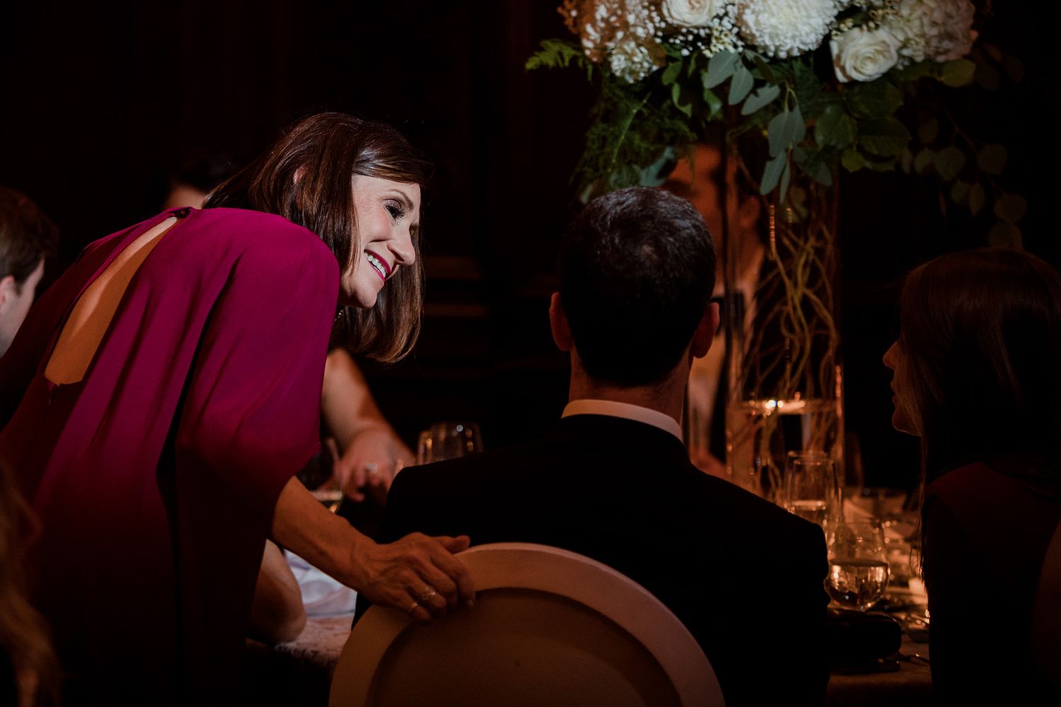 A woman in a red dress leans over to speak to a man seated at a table decorated with tall flowers during an elegant dinner.