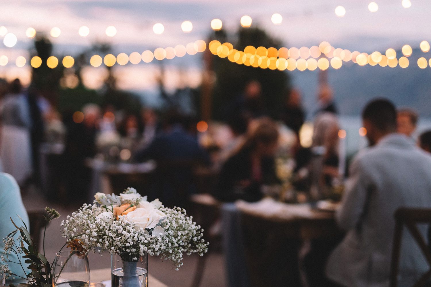 String lights illuminate an outdoor evening wedding reception with guests mingling under a twilight sky.
