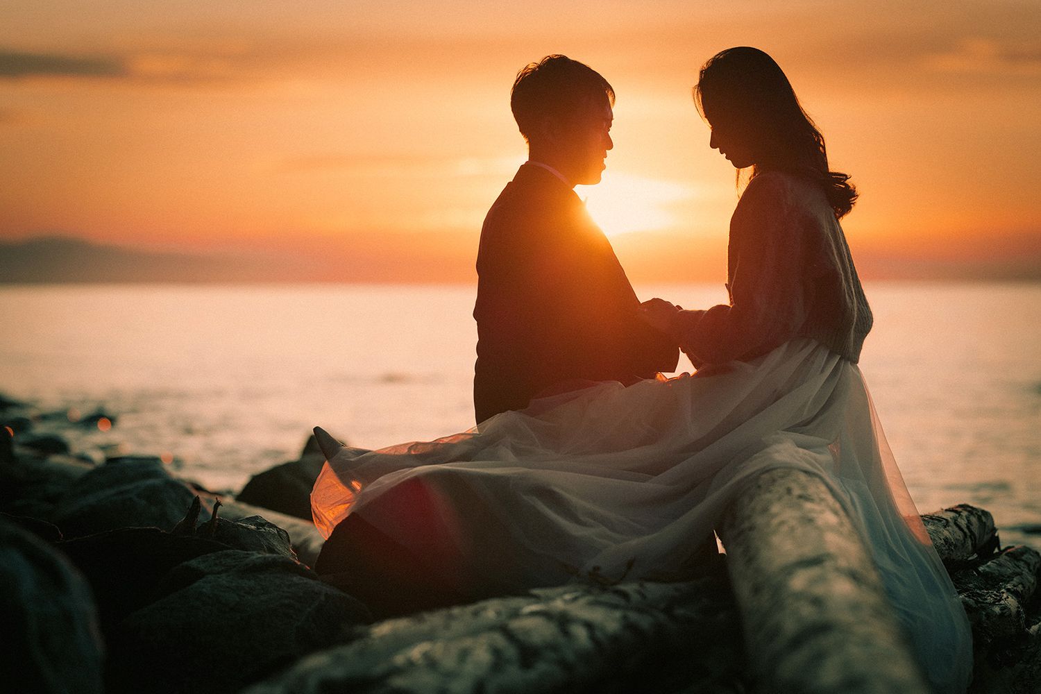 Silhouetted couple embraces on rocky beach at sunset with golden sun and ocean in background.