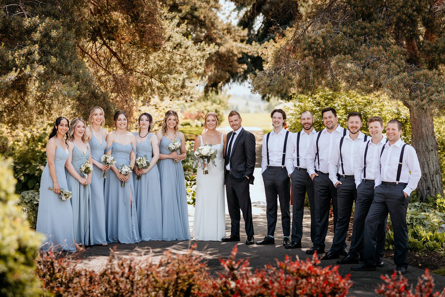 Wedding party in light blue dresses and dark suits poses outdoors under autumn trees for formal group photos.