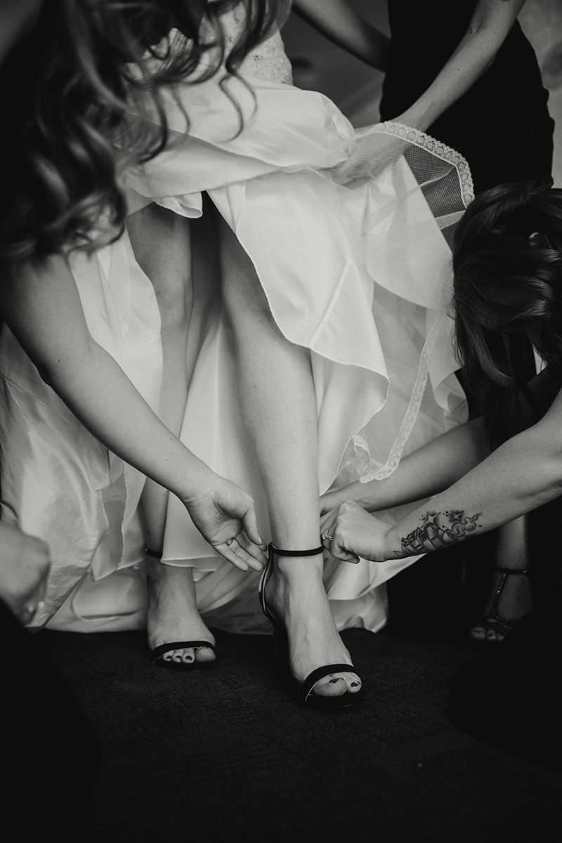 Black and white photo of hands helping to fasten shoes on feet wearing a flowing white wedding dress.