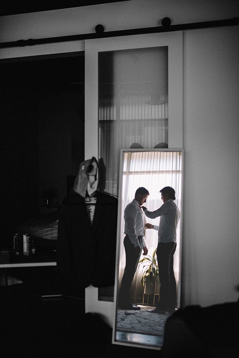 Two men stand in a doorway, reflected in a mirror with a suit jacket hanging in the foreground.