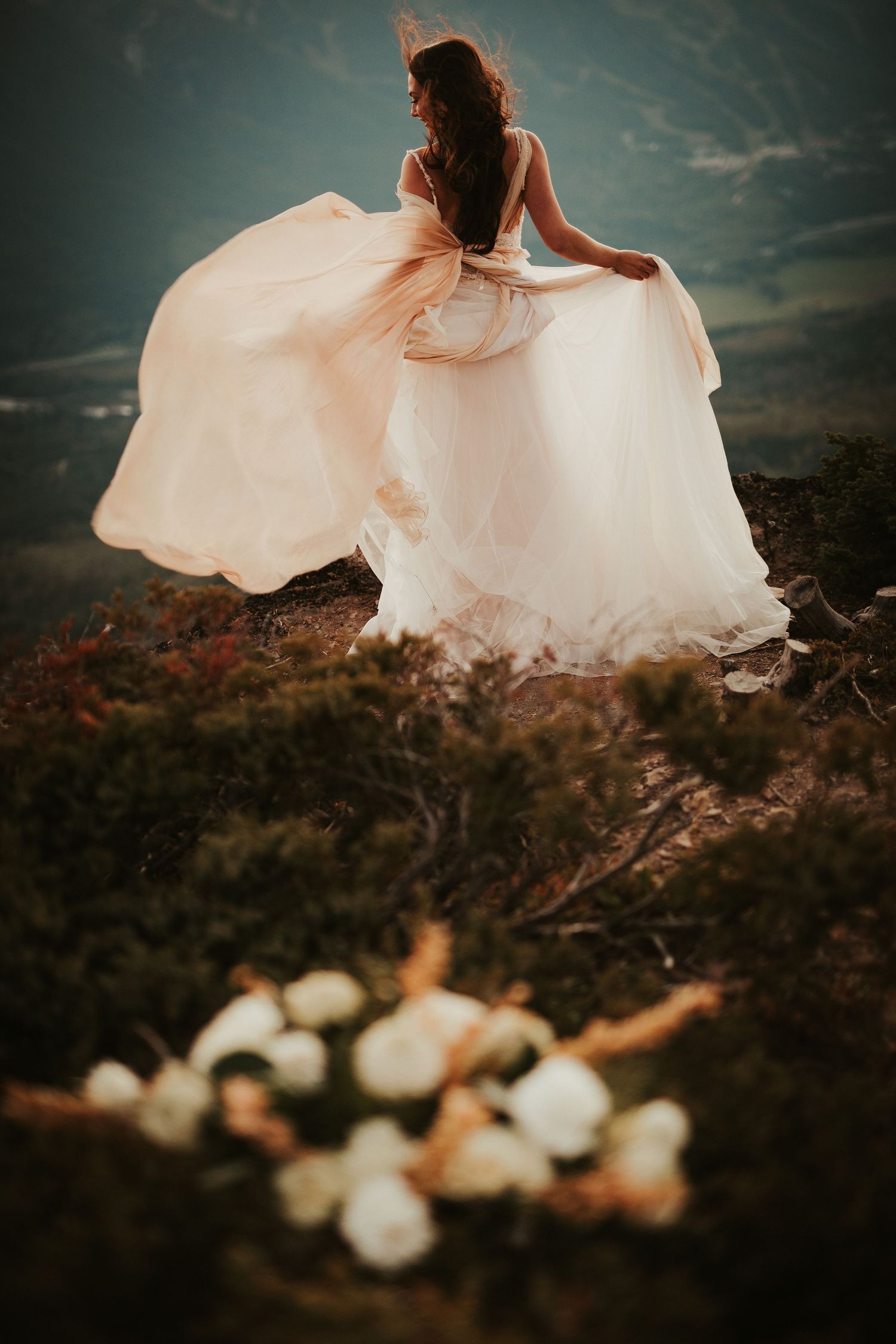 A flowing white wedding dress billows in the wind against a moody sky during an outdoor mountain photo shoot.