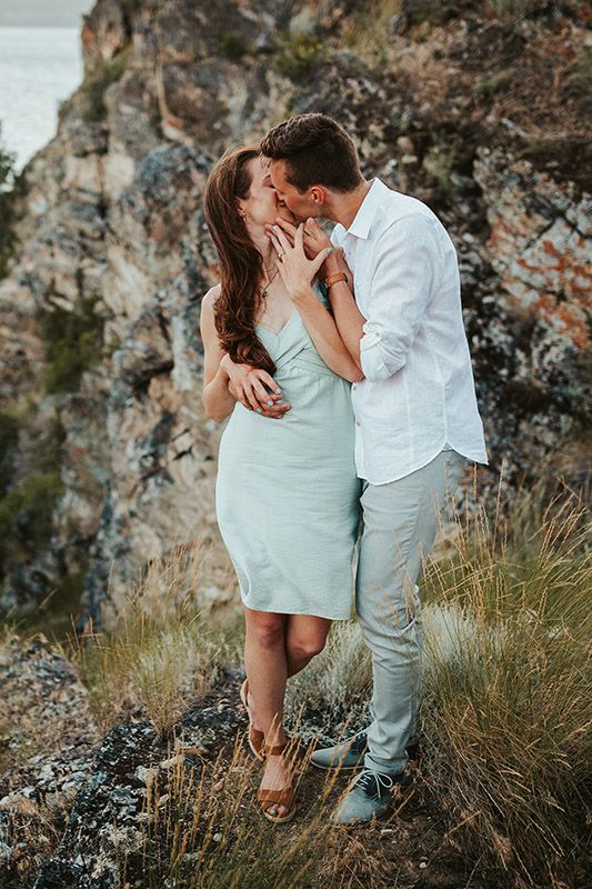 A couple shares a romantic kiss on a rocky hillside, dressed in light summer attire with ocean views behind them.